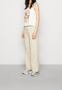 Top bianco senza maniche con stampa grafica, abbinato a pantaloni beige a gamba larga e scarpe da ginnastica grigie con accenti argentati.