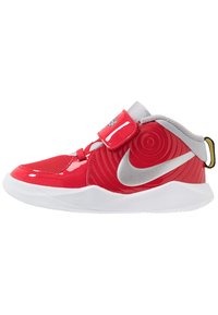 Červená syntetická sportovní bota s lesklým povrchem, šedým jazykem, strukturovanými akcenty, stříbrným logem a bílou podrážkou. Nastavitelný pásek pro přizpůsobení.