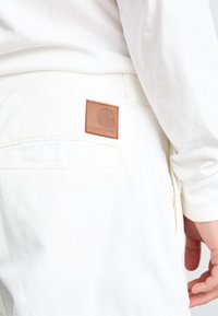 Pantalons blancs avec une pièce en cuir présentant un logo circulaire sur la poche arrière. Le tissu a une texture lisse, avec des détails de couture subtils.