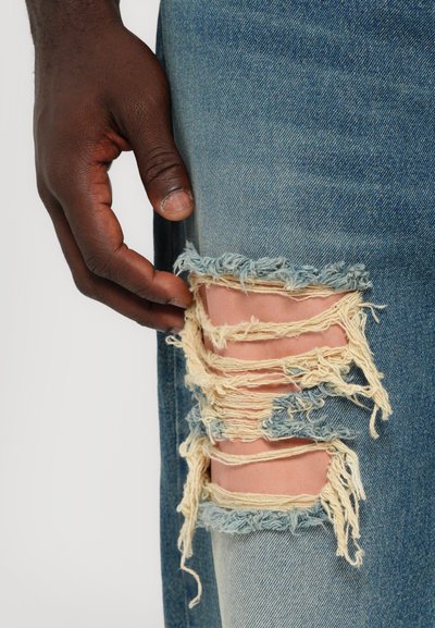Versleten denim jeans met meerdere gescheurde delen. Blauwe stof toont het beige materiaal eronder. Hand die de stof bij de knie aanraakt.
