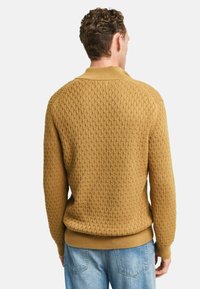 Maglione lavorato a maglia di colore senape con un motivo a diamante testurizzato. Presenta un colletto rotondo e un orlo a costine. Indossato con jeans azzurri chiari.