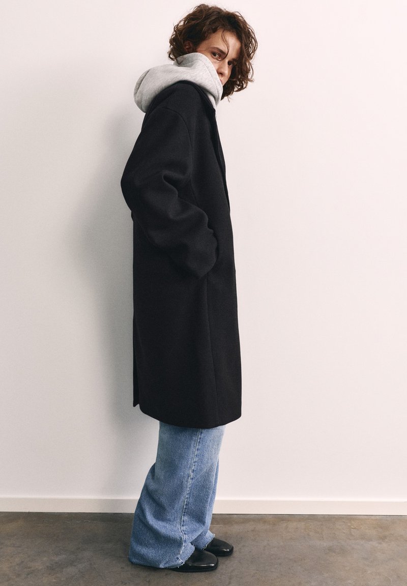 Personne aux cheveux bouclés portant un manteau noir sur un sweat à capuche gris, un jean bleu et des chaussures noires, debout de profil contre un mur blanc.