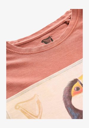 Vaaleanpunainen puuvillapaita, jossa on pyöreä kaula-aukko, ja jossa on painettu kuvasto tucanista ja harp-logosta tarrassa.