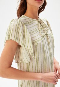 Kevyt raidallinen mekko kermansävyssä, jossa vihreät viivat. Ominaisuuksiin kuuluvat flotterihihat ja nyöriäytteinen kaula-aukko. Pehmeä kangas, rento istuvuus.