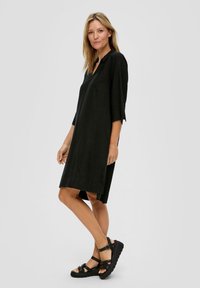 Zwarte jurk tot de knie met een kraag en driekwartmouwen; heeft een relaxed fit en uitsparingen aan de zijkanten. G gedragen met zwarte sandalen met bandjes.