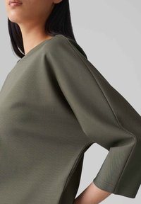 Olijfgroene, lichtgewicht top met een relaxte pasvorm, brede mouwen en een ronde hals. Glad materiaal met zichtbare stikseldetails.