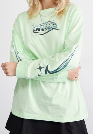 Osoba ubrana w jasnozieloną koszulkę z długim rękawem z stylizowanym napisem „NIKE” i logo typu swoosh z błyszczącymi akcentami na rękawach i klatce piersiowej.