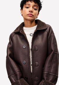 Personne portant une veste en cuir doublée de shearling marron foncé sur une chemise blanche, avec des cheveux courts et bouclés, et des boucles d'oreilles en or.