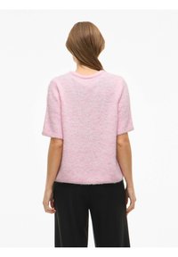 Lühikeste varrukatega roosa pehme kampsun ümarate kaelusega ja pehme tekstuuriga, vaadatuna tagant, mustade pükstega kombineeritud.