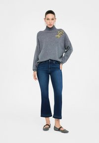 Vrouw staat gekleed in een grijze coltrui met gouden bloemenborduursels, gecombineerd met cropped donkerblauwe spijkerbroeken en zwarte flats.