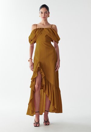 Dlhé šaty bez ramienok v horčicovej žltej farbe s volánovým lemom a predným rozparkom, vyrobené z ľahkej tkaniny. Skombinované s čiernymi sandálmi na opätku.