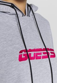 Felpa grigia realizzata in un tessuto morbido, con cappuccio regolabile con dettagli neri e un grande logo "GUESS" ricamato in rosa sul davanti.