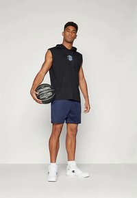 Fekete ujjatlan kapucnis felső logóval, tengerészkék sportnadrág, fekete Spalding kosárlabdát tartva, fehér sportcipőt viselve. Egyszerű, sportos dizájn.