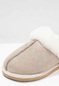 Pantofole in suede beige con polsino in pelliccia bianca, caratterizzate da una cucitura lungo la parte superiore e una suola in gomma di colore chiaro.