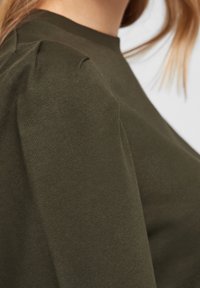 Ciemnozielona bluzka z długim rękawem i okrągłym dekoltem, z teksturowanym materiałem i marszczeniem na ramieniu. Zbliżenie na materiał.