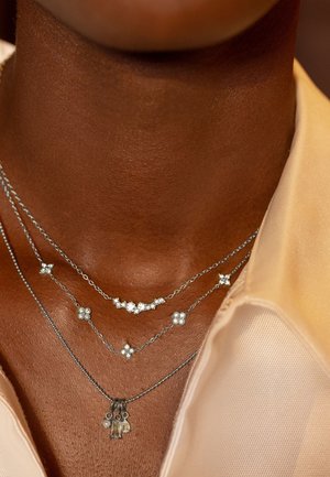 Trois colliers en argent superposés sur une peau foncée avec des pendentifs en forme de losange et en grappe, portés sur une chemise beige à col.