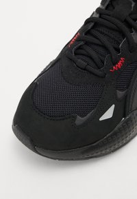 Black sneaker featuring a mesh upper and suede accents, textured sole, and red stitching details. Includes a reflective silver accent.