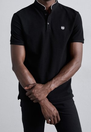 Schwarzes Polohemd mit Stehkragen, weißem Besatz und kleinem gesticktem Logo auf der Brust. Der Stoff wirkt glatt und enganliegend.