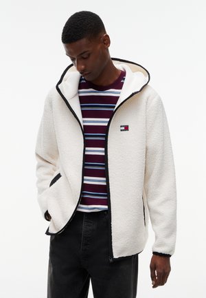 Joven con chaqueta de forro polar blanca con capucha, camisa a rayas en burdeos y azul, y pantalones negros, mirando hacia abajo con una mano en el bolsillo.