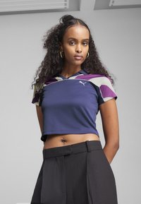 Violetinė ir tamsiai mėlyna trumpa sportinė marškinėlė trumpomis rankovėmis, su trikampio formos akcentais, Puma logotipas ant krūtinės, derinama su aukšto liemens juodomis kelnėmis.