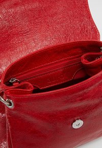 Sac bandoulière en cuir rouge avec une finition texturée, compartiment zippé et ferrures argentées. Comprend une poche intérieure et une fermeture magnétique.