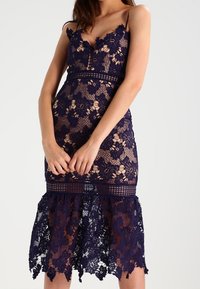 Námořnický modrý krajkový šat s květinovým vzorem, upnutým korzetem a rozšířeným lemem; má nastavitelné ramínka a průhlednou strukturu.