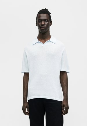 Čierny muž s dredmi, oblečený v bielej polokošeli s krátkym rukávom a čiernych nohaviciach, stojaci pred jednoduchým svetlým pozadím.