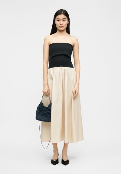 Γυναίκα που φοράει μαύρο τοπ χωρίς τιράντες και φούστα σε κρεμ απόχρωση, κρατώντας μια μαύρη καπιτονέ τσάντα με αλυσίδα, στέκεται μπροστά σε ένα απλό φόντο.