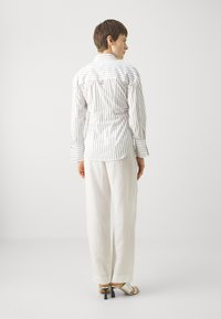 Hvitstripet skjorte med lange ermer, svart med lyse beige linbukser og åpne hæler, sett fra baksiden.