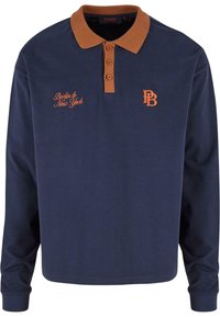 Mørkeblå langermet polo skjorte med brun krage, med tre knapper og oransje broderi som leser "Berlin til New York" og initialene "PB."