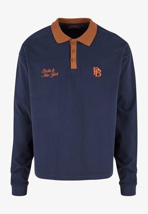 Tengerészkék, hosszú ujjú pólóing, barna gallérral, három gombbal, narancssárga hímzéssel, amelyen a "Berlin to New York" felirat és a "PB" kezdeti betűk találhatók.