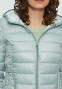 Chaqueta acolchada de color azul claro con costuras horizontales, cuello alto y cremallera frontal completa. Presenta una capucha y una textura suave.