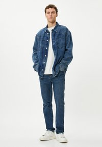 Chaqueta de mezclilla azul oscura con grandes bolsillos frontales, camiseta blanca debajo, jeans azules ajustados y zapatillas blancas. Estilo clásico y casual.