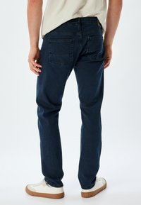 Jeans de mezclilla azul oscuro con un corte ajustado, que cuentan con un diseño clásico de cinco bolsillos y detalles de costura sutiles. Combinados con zapatillas blancas.