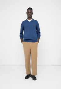 Kék V-nyakú pulóver kis sárga logóval, bézs kordbársony nadrág, és fekete cipők. A modell semleges háttér előtt áll.