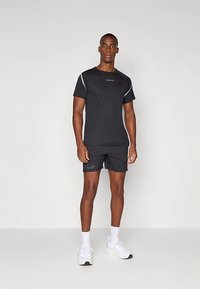 Vyras, dėvintis juodą sportinę marškinėlę ir šortus, baltas kojines ir baltus sportbačius, stovintis priešais paprastą šviesų foną.