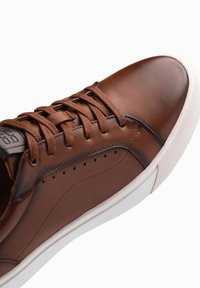 Baskets en cuir marron avec bout rond, lacets plats, détails perforés sur le côté et semelle en caoutchouc blanche. Texture lisse et design minimal.