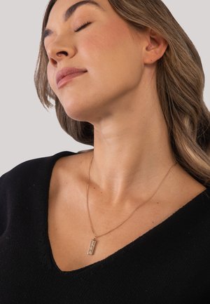 Frau mit geschlossenen Augen, die den Kopf leicht zurückneigt und ein schwarzes Oberteil sowie eine Halskette mit einem goldenen Stabanhänger trägt.