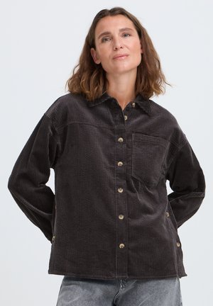Femme aux cheveux bruns mi-longs porte une chemise ample en velours côtelé marron foncé à boutons et un jean gris clair, debout devant un fond uni.