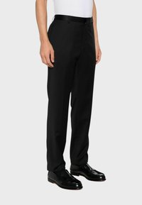 Pantaloni formali neri realizzati in tessuto liscio, con vestibilità su misura, caratterizzati da una parte anteriore piatta, tasche laterali e una cintura elegante. Indossati con mocassini neri.
