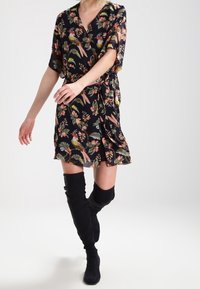 Kvinna klädd i en svart blommig klänning med fåglar och knähöga svarta mockastövlar som går mot en enfärgad bakgrund.