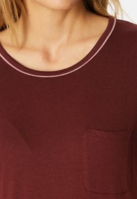 Mujer vistiendo una camiseta color burdeos con un pequeño bolsillo en el pecho y un cuello redondo delineado con un ribete de color rosa claro.