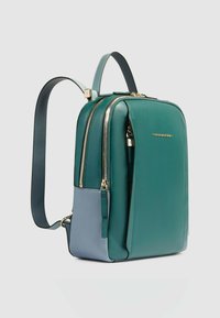 Mochila de cuero azul verdoso con textura suave, compartimentos dobles con cremallera, detalles de herrajes dorados y una base de color azul claro en contraste.