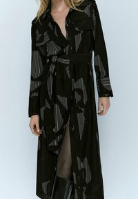 Abito lungo nero con maniche a righe, bottoni frontali, vita con cintura e due tasche applicate sul petto.