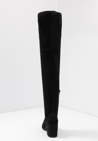 Sort ruskind støvle over knæet med blokhæl, udstyret med lynlås på ydersiden og lodrette sømdetaljer for struktur.