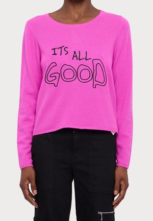 Jasnoróżowy sweter z długim rękawem z czarnym napisem "ITS ALL GOOD" noszony przez osobę w czarnych spodniach na jednolitym tle.