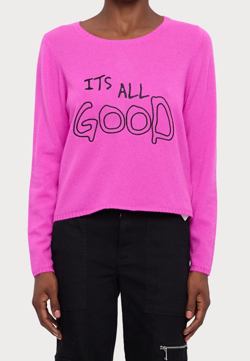 Pull rose vif à manches longues avec le texte noir « ITS ALL GOOD » porté par une personne en pantalon noir sur un fond uni.