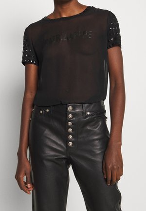 Czarny przezroczysty top z krótkim rękawem z ćwiekami na rękawach i wytłoczonym logo, zestawiony z czarnymi skórzanymi spodniami z wysokim stanem i srebrnymi guzikami.