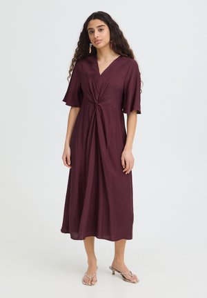 Hosszú, göndör hajú nő, aki egy szilvaszínű, midi hosszúságú, rövid ujjú, derékrésznél csomós részlettel ellátott ruhát visel, amelyet magassarkú szandállal kombinál.