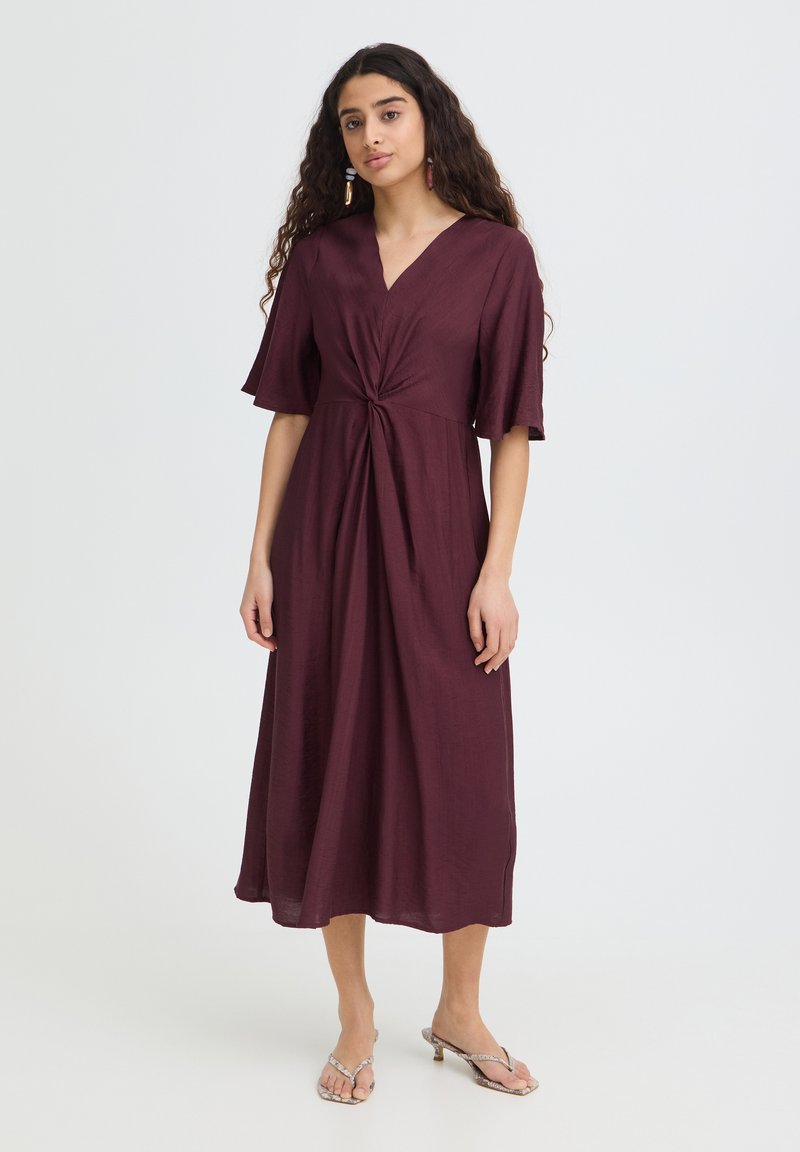 Femme aux longs cheveux bouclés portant une robe midi prune à manches courtes avec un détail noué à la taille, assortie à des sandales à talons.